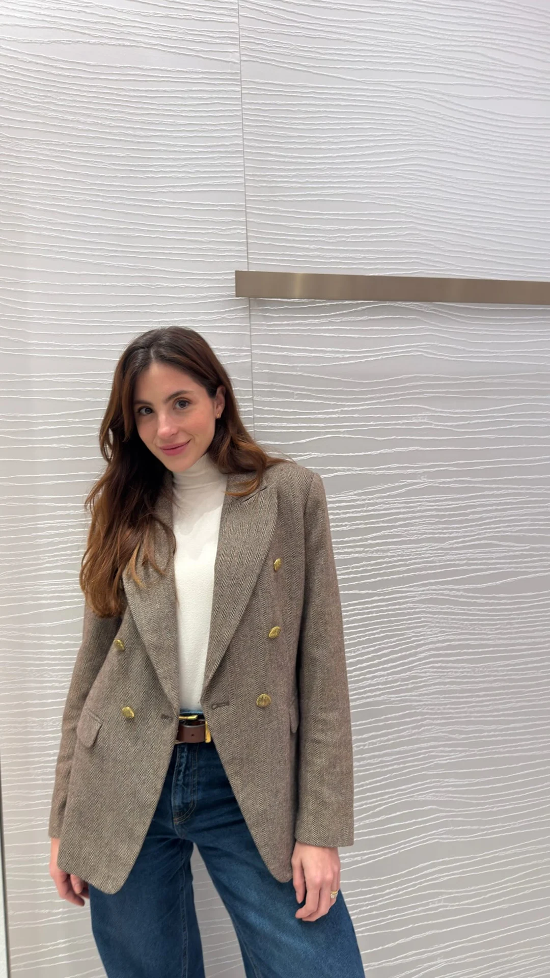 Rafaela de blazer marrom e jeans, diante de uma parede clara